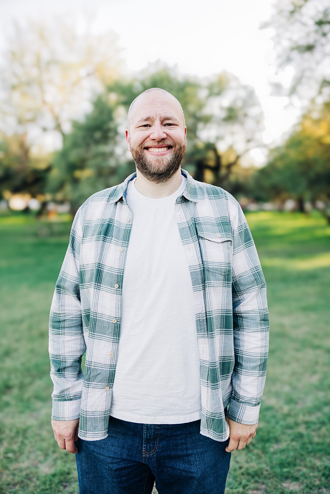 Smiling man in a green and white plaid shirt over a white t-shirt and blue jeans, standing in a sunlit park with green grass and trees, captured on a bright, clear day.