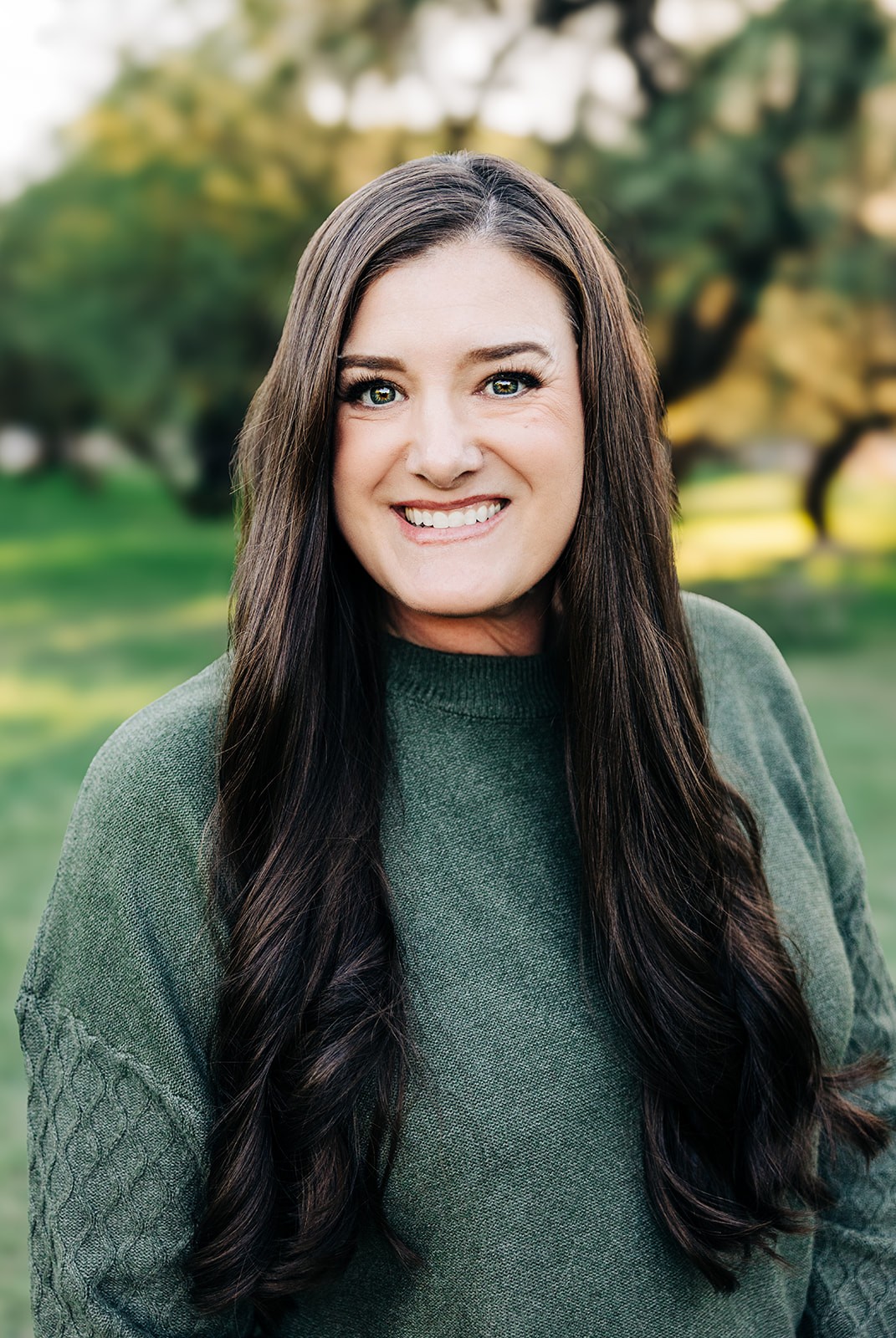 Woman with long dark brown hair in a green textured sweater standing in a sunlit park with trees and grass, captured during golden hour for a warm, natural aesthetic.