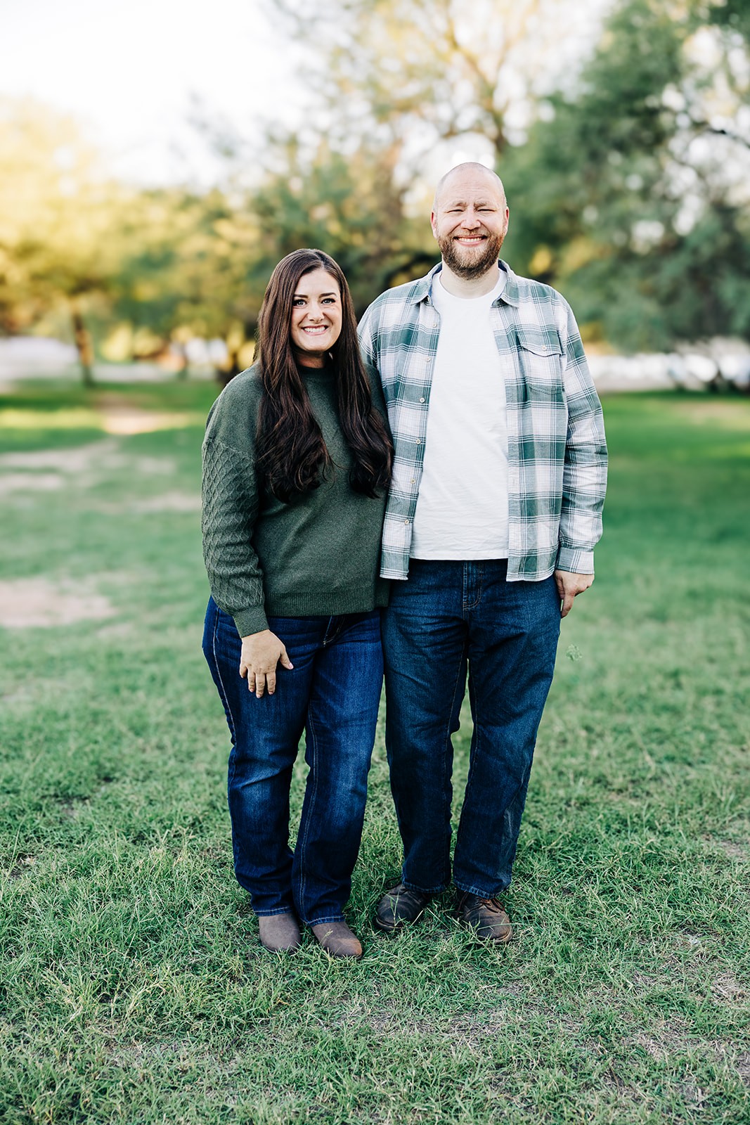 Smiling couple standing side by side in a sunlit park, wearing casual outfits—green sweater and jeans for her, plaid shirt and jeans for him—surrounded by trees and grass during golden hour.
